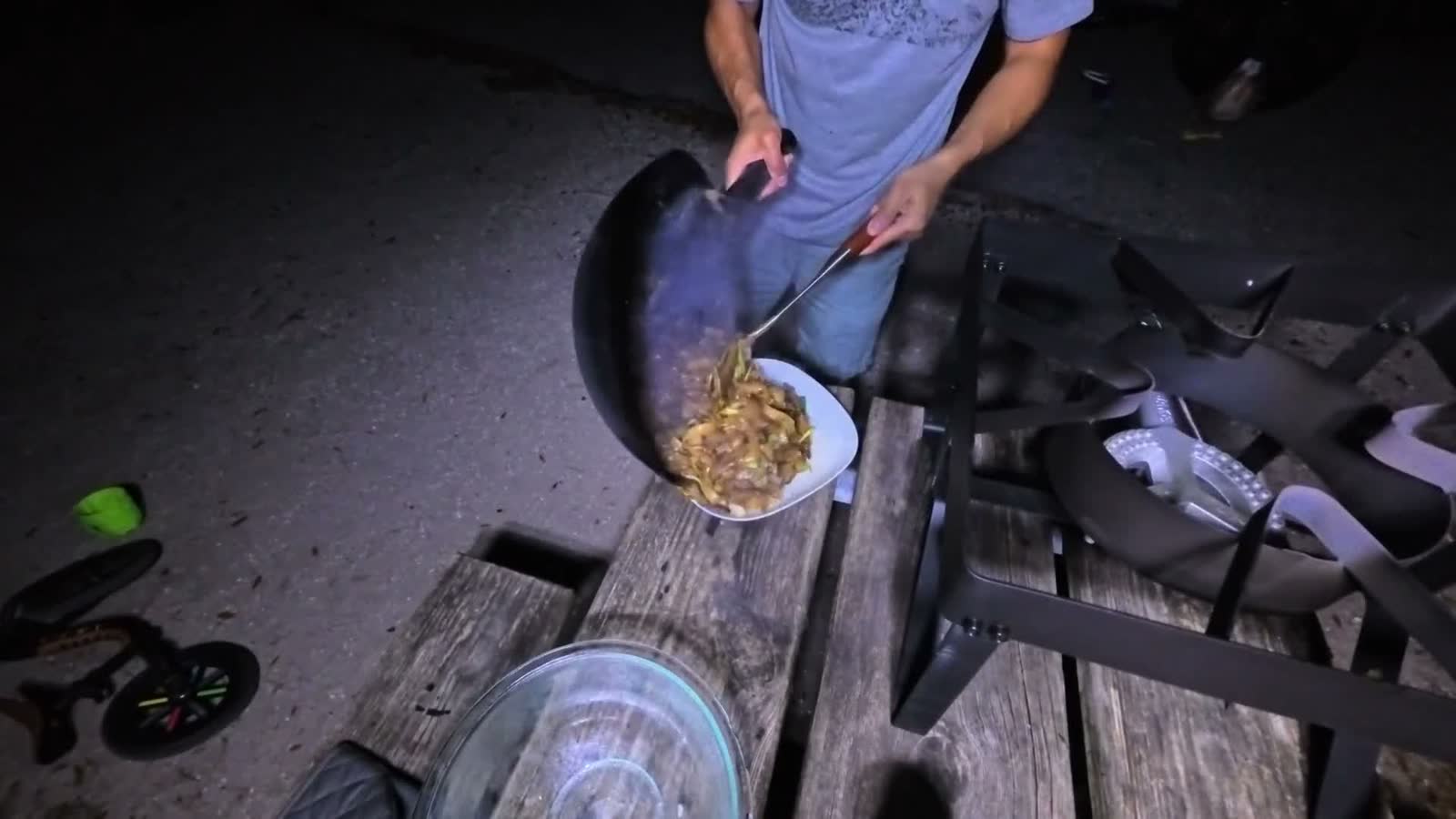 Michael sliding beef chow fun from a wok onto a plate at a picnic table at night