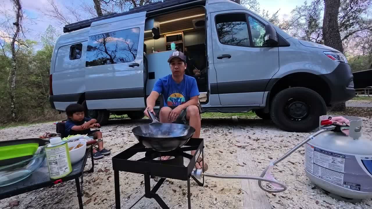 Michael at a wok on a propane burner beside the open side door of the van, with Spencer sitting nearby