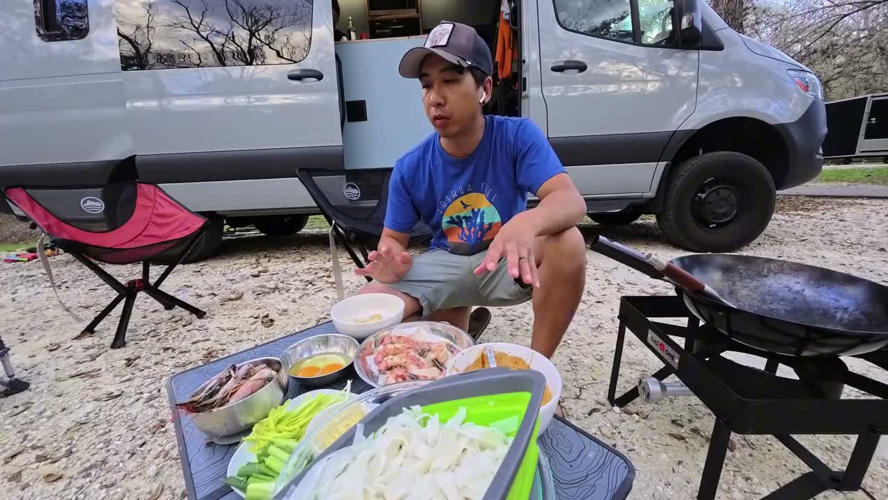 Michael crouched next to a table of prepped shrimp, eggs, noodles and greens beside a wok on a propane burner outside the van