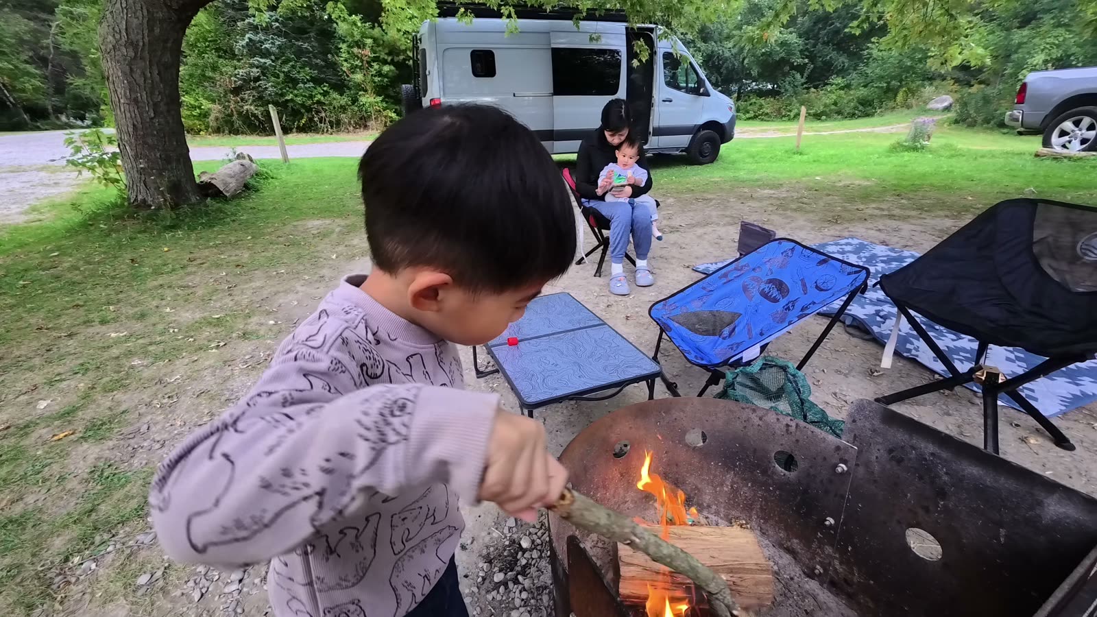 Spencer adding a stick to the campfire while Rina holds Archie in a camping chair next to our van
