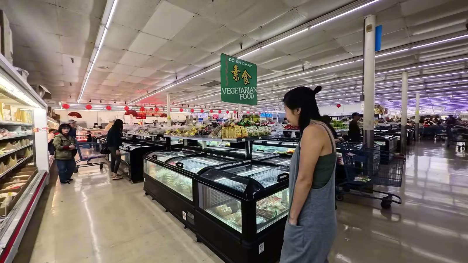 Rina walking past freezer cases under a Vegetarian Food sign in a large Asian supermarket