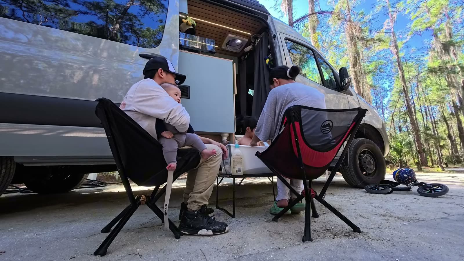 Michael holding Archie in a camp chair while Rina bathes Spencer in a foldable tub outside the van at a Florida campground