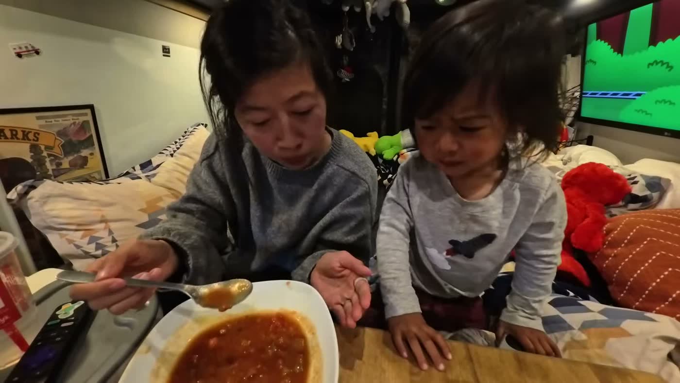 Rina giving Spencer a spoonful of borscht from a bowl at the table in the van