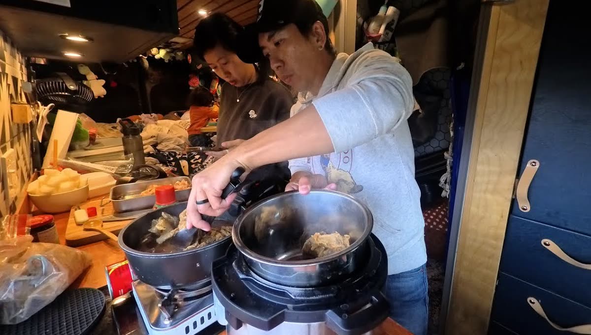 Michael lifting blanched beef from a pan into a small pressure cooker in the van kitchen, with Rina beside him