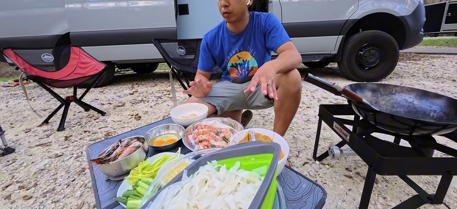 Michael sitting next to the wok burner at the campground with bowls of shrimp, noodles, greens and sauce laid out on a small table