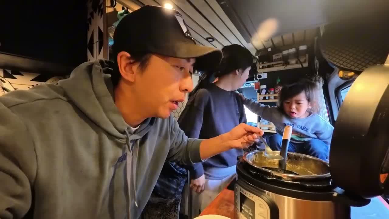 Michael tasting the soup from the pot while Rina and Spencer stand next to him in the van kitchen
