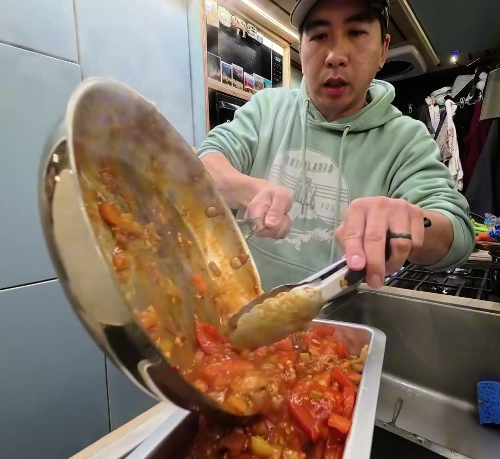 Michael pouring tomato sauce with vegetables from a pan into a baking pan of spaghetti at the van sink