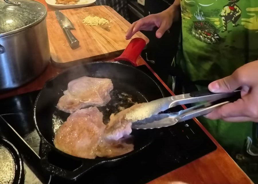 Thin pork chops frying in a cast iron pan, flipped with tongs, chopped garlic on the cutting board behind