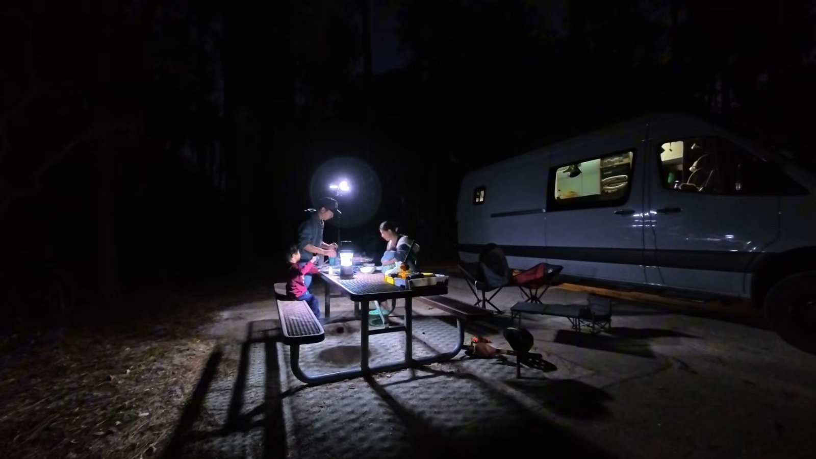 Our family at a campground picnic table at night, lit by a camping light, with the van parked next to the site