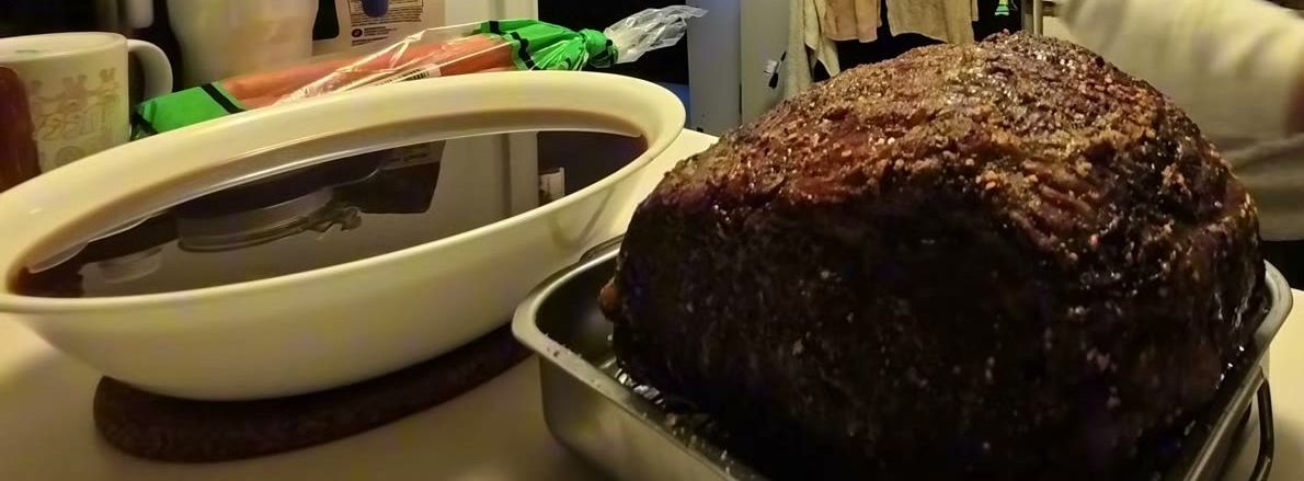 A browned prime rib roast just out of the oven on a wire rack, next to a big bowl of dark au jus