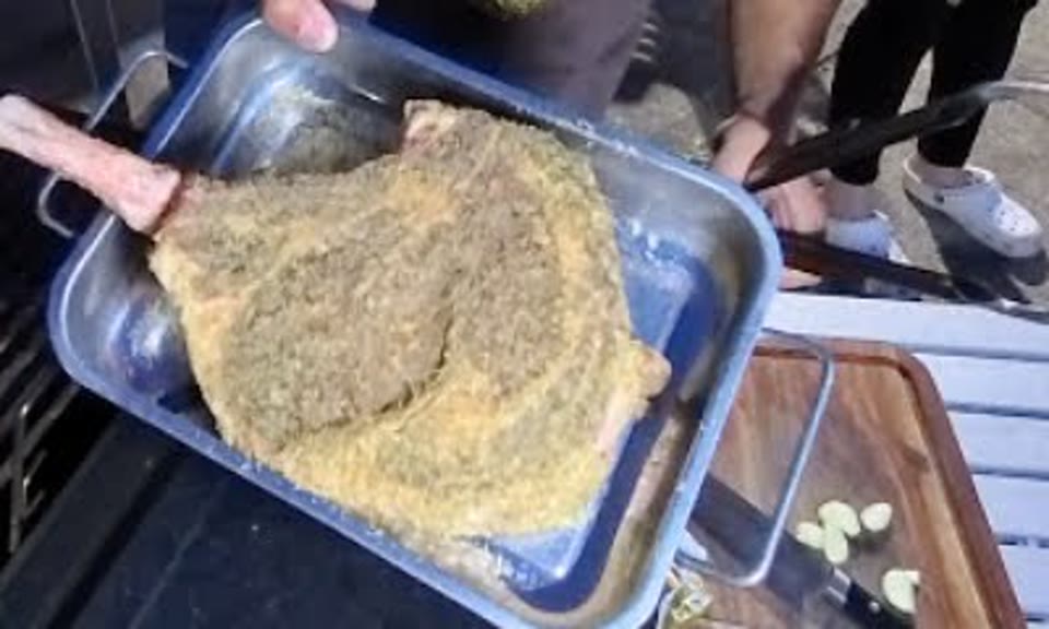 A raw tomahawk steak covered in seasoning in a metal tray next to the grill, with garlic cloves on a cutting board